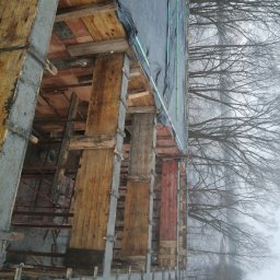 Usługi budowlane Kamil Krupinski - Wznoszenie betonowego szkieletu budynku z drewnianym szalunkiem, widok na fundamenty i konstrukcję w surowym stanie, z drzewami w tle.