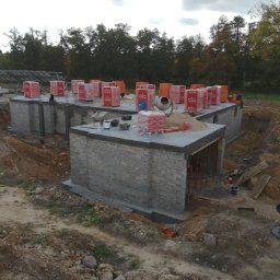 Usługi budowlane Kamil Krupinski - Wznoszenie ścian parteru z bloczków ceramicznych na betonowym fundamencie, widoczne palety z bloczkami, betoniarka i wykop ziemny wokół budynku.