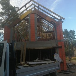 Usługi budowlane Kamil Krupinski - Wznoszony murowany dom z czerwonej cegły, widoczna konstrukcja dachu z drewnianych belek, częściowo zasłonięty przez samochód dostawczy z logiem Jegger, w tle drzewa i błękitne niebo.