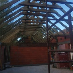 Usługi budowlane Kamil Krupinski - Wnętrze domu w trakcie budowy: widoczne ściany z czerwonej cegły Porotherm, drewniana konstrukcja dachu i rusztowanie. Materiały budowlane składowane wewnątrz. Niebo widoczne przez konstrukcję dachu.