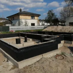 Zabezpieczony fundament domu jednorodzinnego z wystającymi prętami zbrojeniowymi, czarna folia izolacyjna, w tle budynki mieszkalne pod błękitnym niebem.
