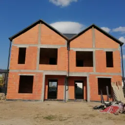 Budowa bliźniaka z czerwonej cegły Porotherm, widoczne otwory okienne i drzwiowe, fundament zabezpieczony czarną izolacją, palety i materiały budowlane na placu budowy.