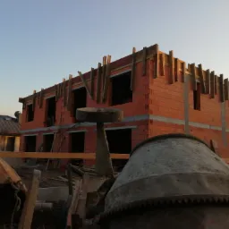 Dwukondygnacyjny budynek w trakcie wznoszenia ścian z czerwonej cegły, widoczne drewniane szalunki stropowe, na pierwszym planie zardzewiała betoniarka i inne narzędzia budowlane, niebo...