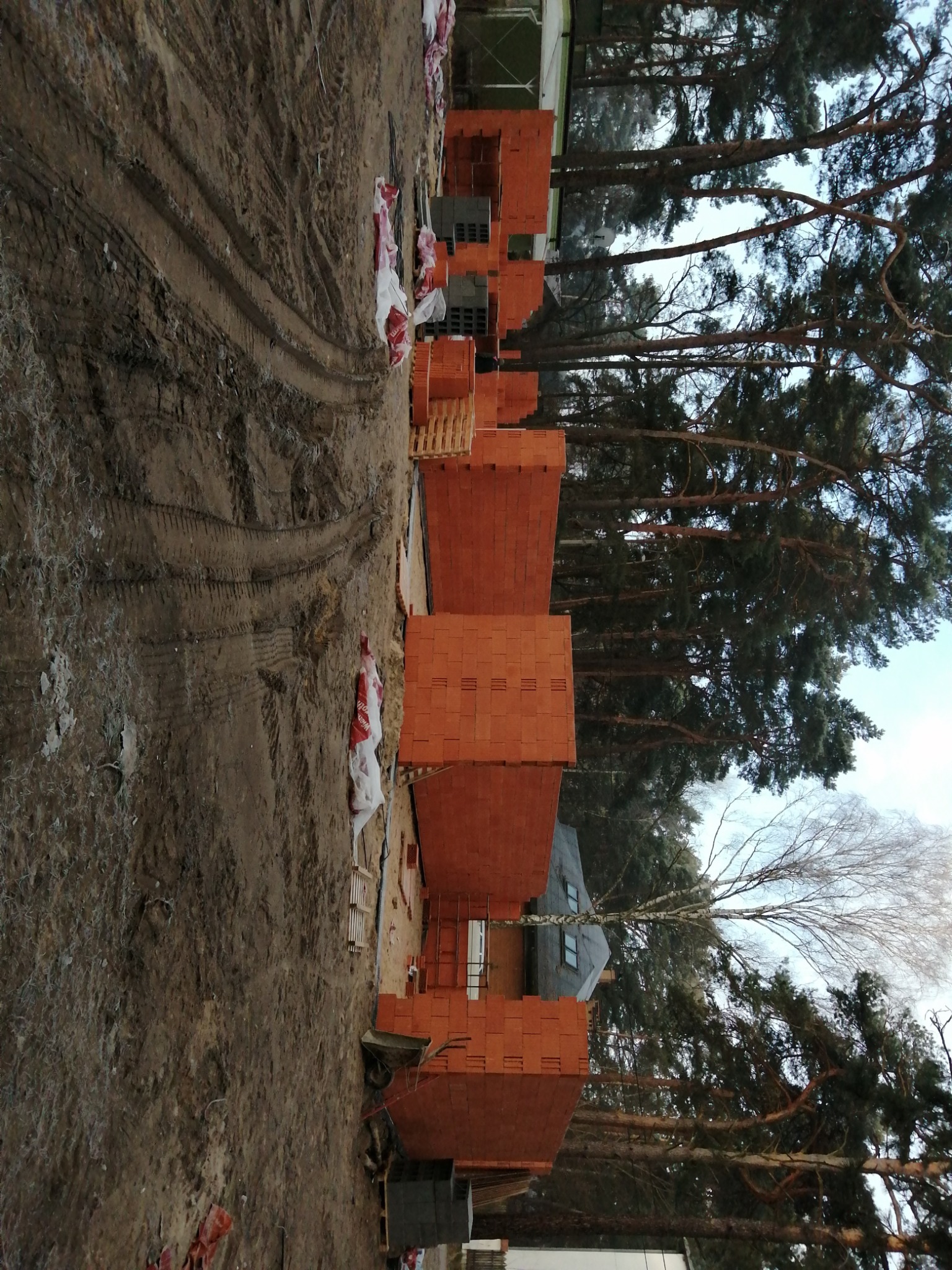 Wznoszone ściany z czerwonej cegły na placu budowy, widoczne palety z cegłami, ślady opon na błotnistym podłożu i wysokie drzewa w tle.