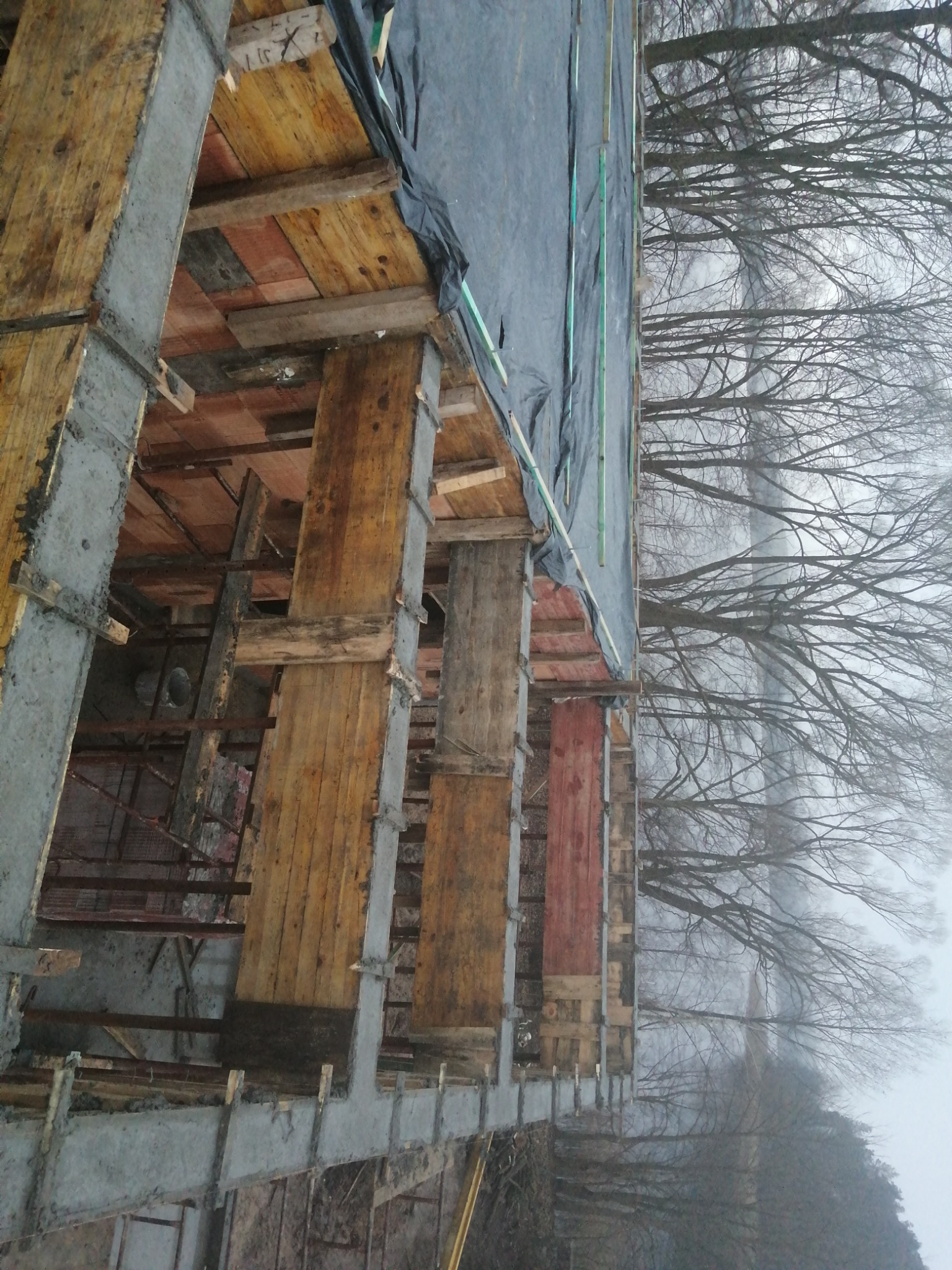 Wznoszenie betonowego szkieletu budynku z drewnianym szalunkiem, widok na fundamenty i konstrukcję w surowym stanie, z drzewami w tle.