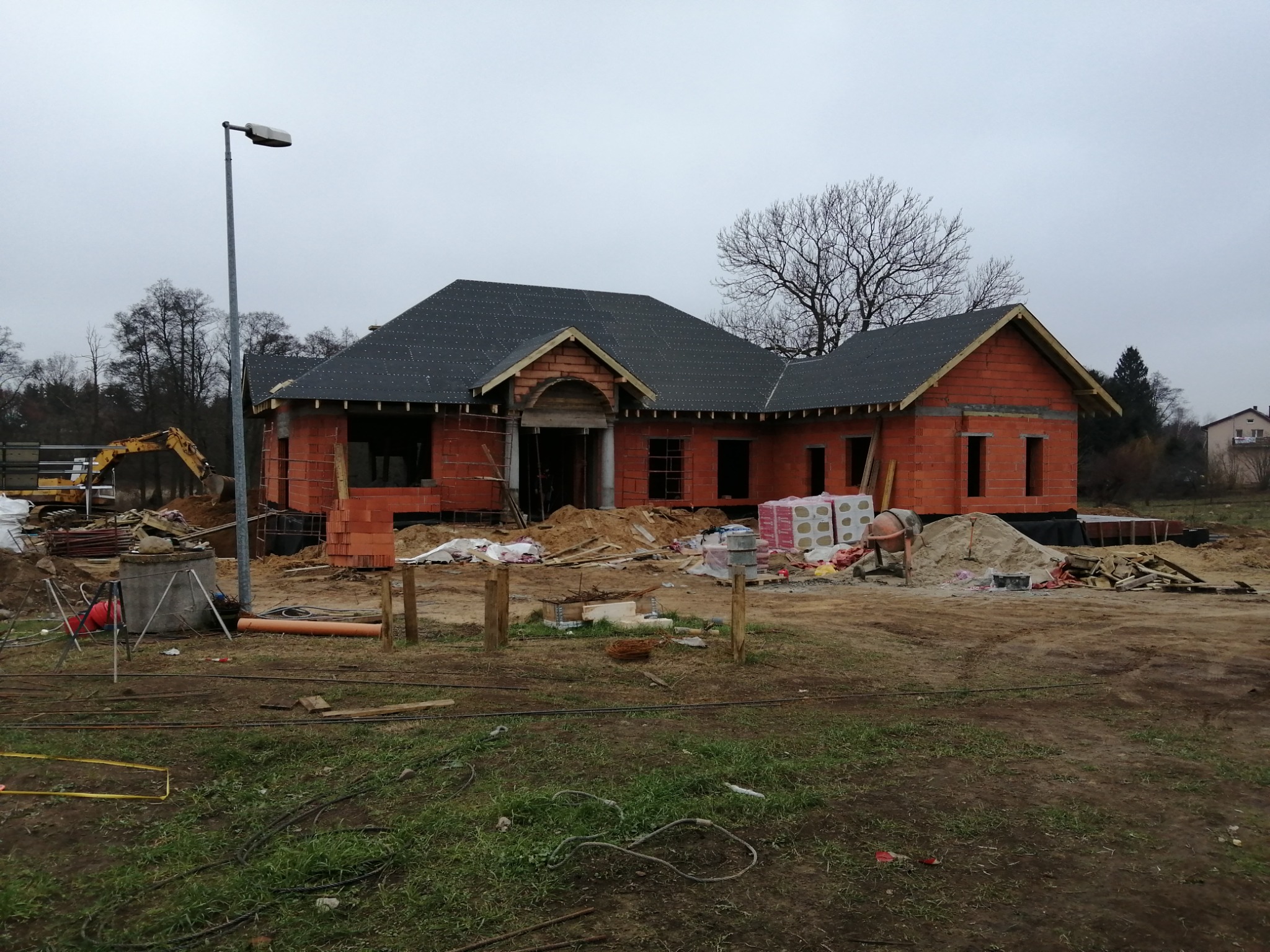 Dom murowany w trakcie budowy z czerwonej cegły, widoczne elementy dachu, teren budowy z ziemią, gruzem i materiałami budowlanymi, w tle drzewa i pochmurne niebo.