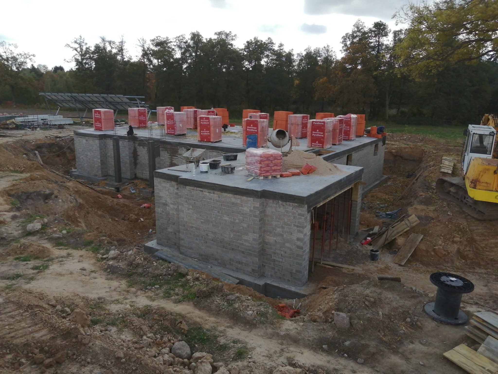 Wznoszenie ścian parteru z bloczków ceramicznych na betonowym fundamencie, widoczne palety z bloczkami, betoniarka i wykop ziemny wokół budynku.