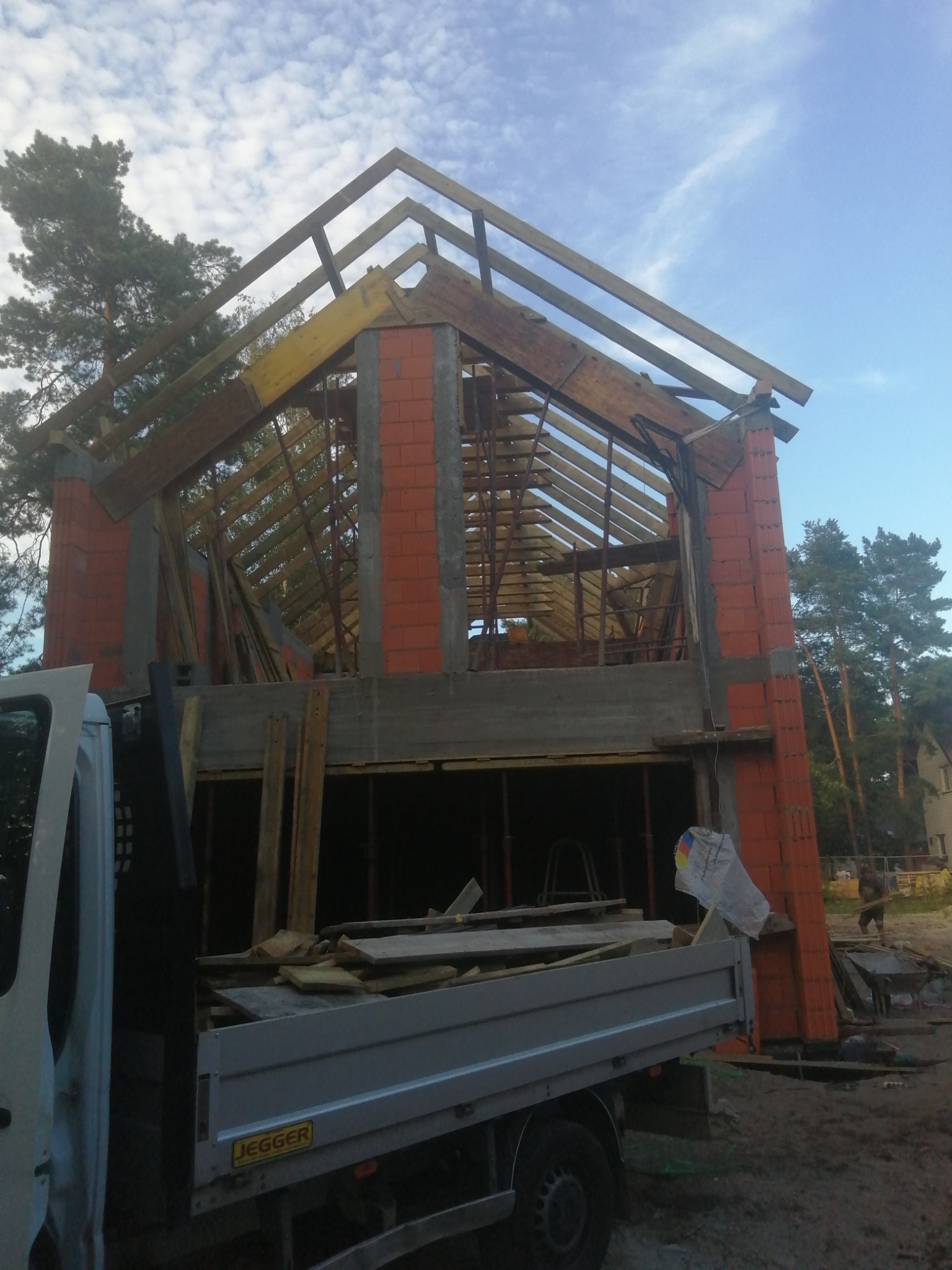 Wznoszony murowany dom z czerwonej cegły, widoczna konstrukcja dachu z drewnianych belek, częściowo zasłonięty przez samochód dostawczy z logiem Jegger, w tle drzewa i błękitne niebo.