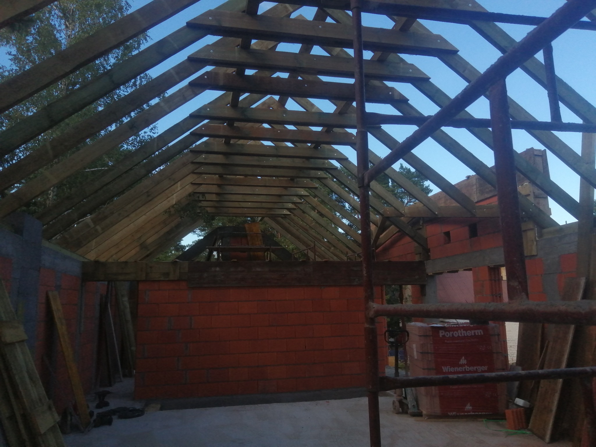 Wnętrze domu w trakcie budowy: widoczne ściany z czerwonej cegły Porotherm, drewniana konstrukcja dachu i rusztowanie. Materiały budowlane składowane wewnątrz. Niebo widoczne przez konstrukcję dachu.