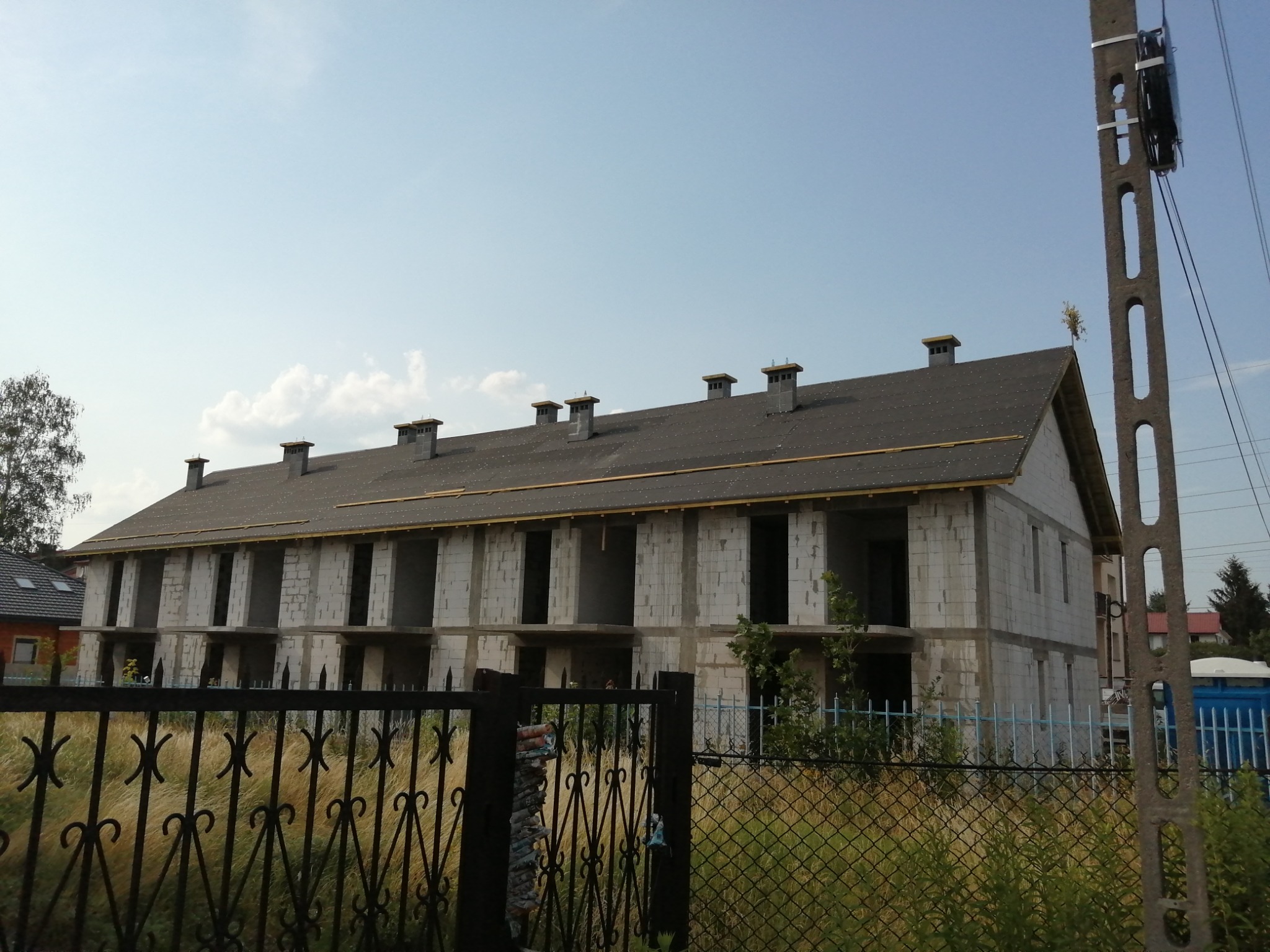 Wielorodzinny budynek w stanie surowym zamkniętym, widoczny zza metalowego ogrodzenia, z kominami i dachem pokrytym papą. Na pierwszym planie słup energetyczny.