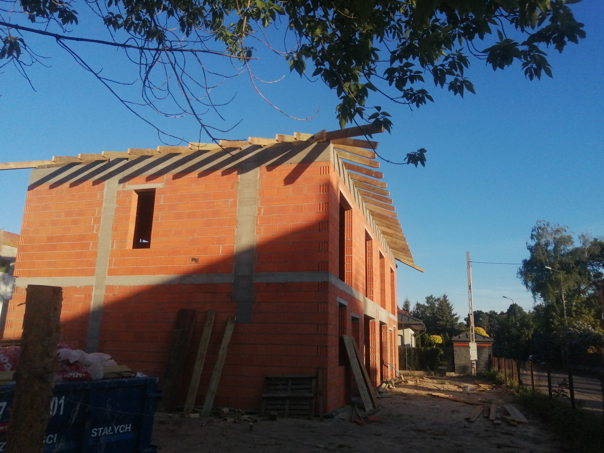 Wznoszony dwukondygnacyjny budynek z czerwonej cegły, z widocznymi elementami szalunku na dachu i niezadaszonymi otworami okiennymi, stojący na piaszczystej działce z niebieskim kontenerem...
