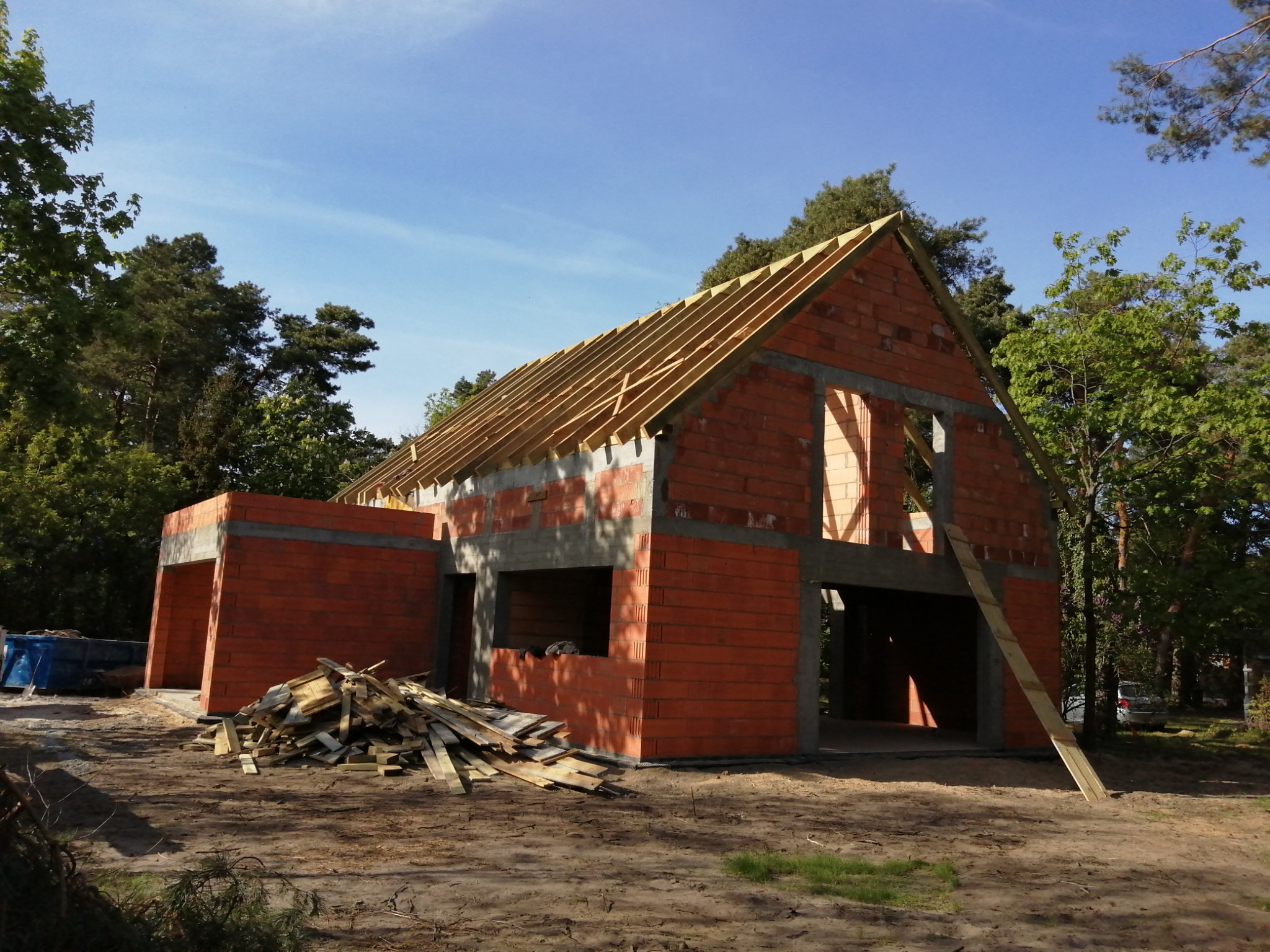 Surowy stan domu z czerwonej cegły z widoczną konstrukcją dachu, drewnianymi belkami i stertą desek przed budynkiem na piaszczystym terenie.