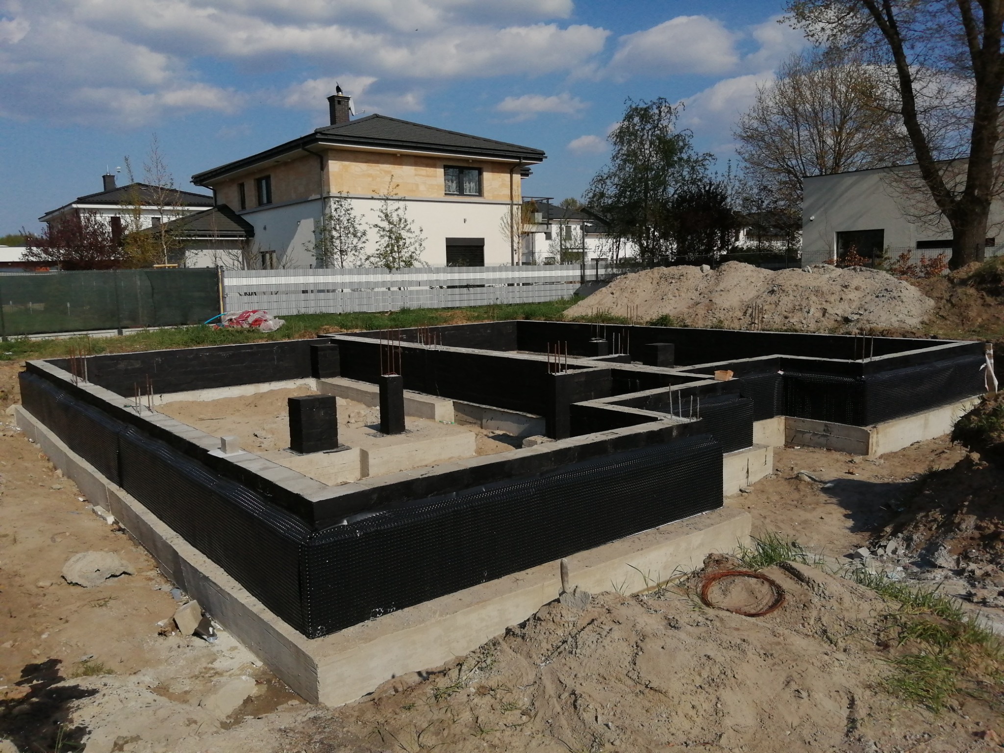 Zabezpieczony fundament domu jednorodzinnego z wystającymi prętami zbrojeniowymi, czarna folia izolacyjna, w tle budynki mieszkalne pod błękitnym niebem.