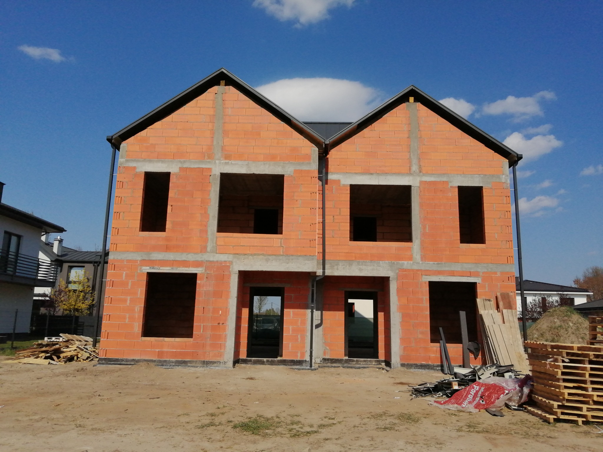 Budowa bliźniaka z czerwonej cegły Porotherm, widoczne otwory okienne i drzwiowe, fundament zabezpieczony czarną izolacją, palety i materiały budowlane na placu budowy.