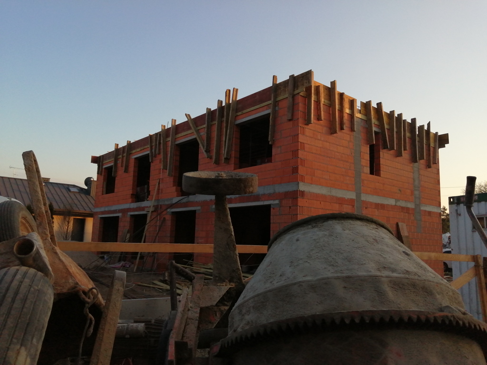 Dwukondygnacyjny budynek w trakcie wznoszenia ścian z czerwonej cegły, widoczne drewniane szalunki stropowe, na pierwszym planie zardzewiała betoniarka i inne narzędzia budowlane, niebo...