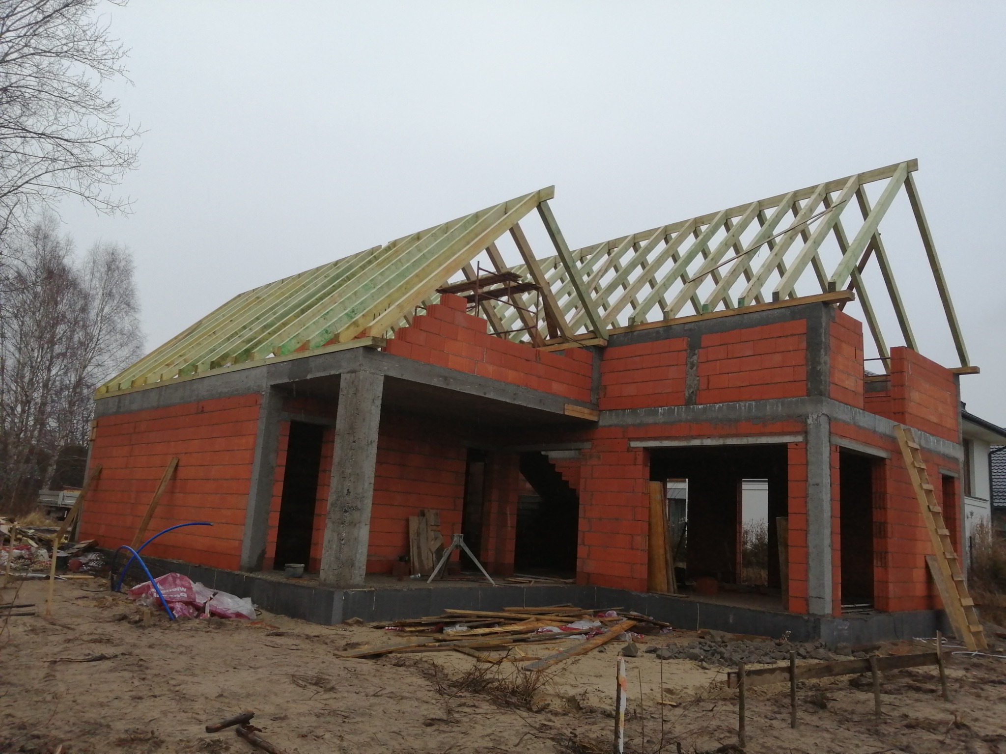 Niedokończony budynek z czerwonej cegły z drewnianą konstrukcją dachu, widoczne fundamenty i ściany, teren budowy z elementami konstrukcyjnymi i narzędziami, pochmurne niebo w tle.