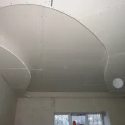 Niedokończony sufit z płyt kartonowo-gipsowych z dekoracyjnymi, falistymi obniżeniami, widoczne łączenia płyt i wkręty, ściany w stanie surowym, okno w tle.