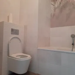 Wykończona łazienka z podwieszaną toaletą, jasnymi płytkami i motywem liści palm na ścianie nad wanną.