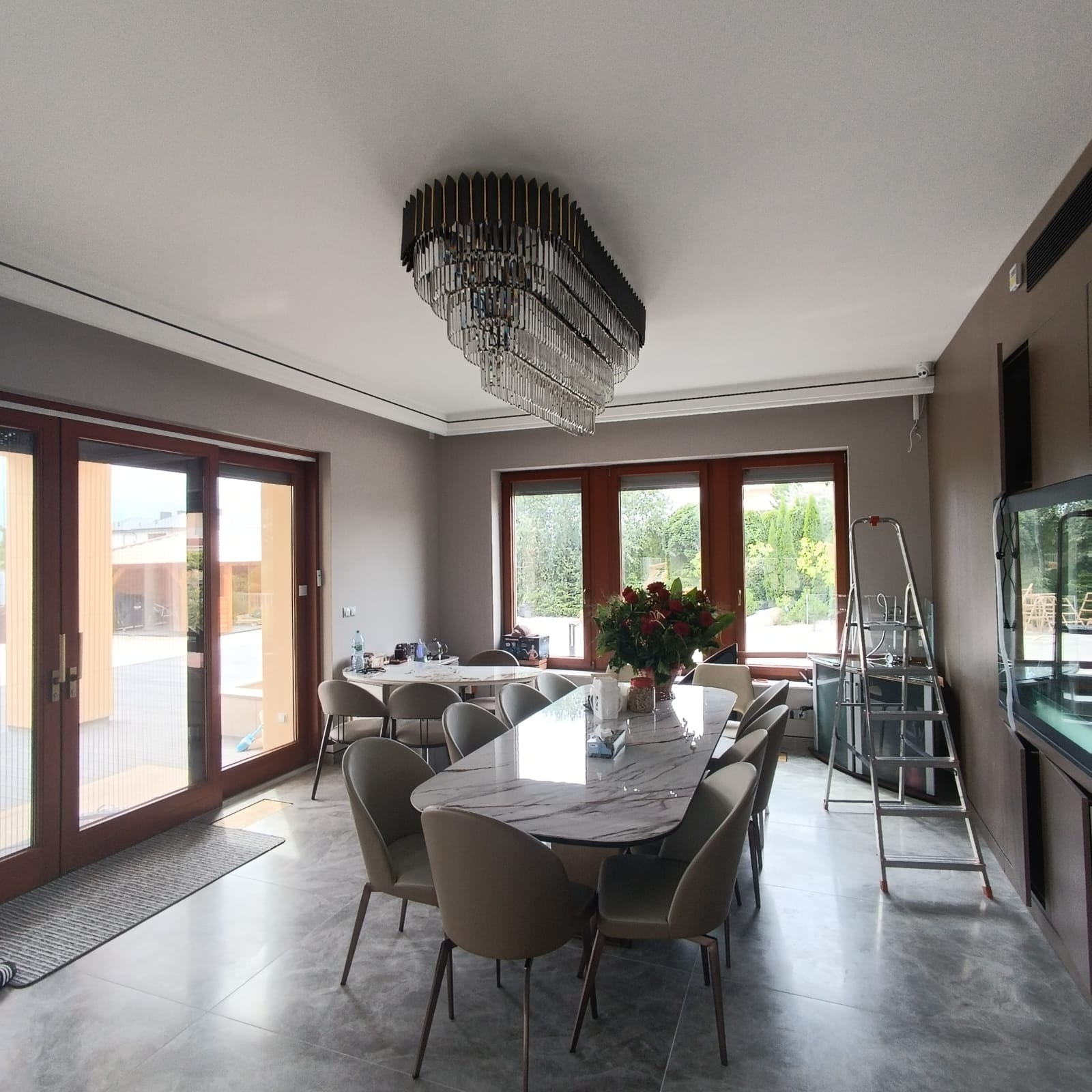Jasne wnętrze jadalni z marmurowym stołem, krzesłami w kolorze taupe, kryształowym żyrandolem i dużymi oknami z widokiem na ogród; drabina oparta o ścianę sugeruje prace wykończeniowe.