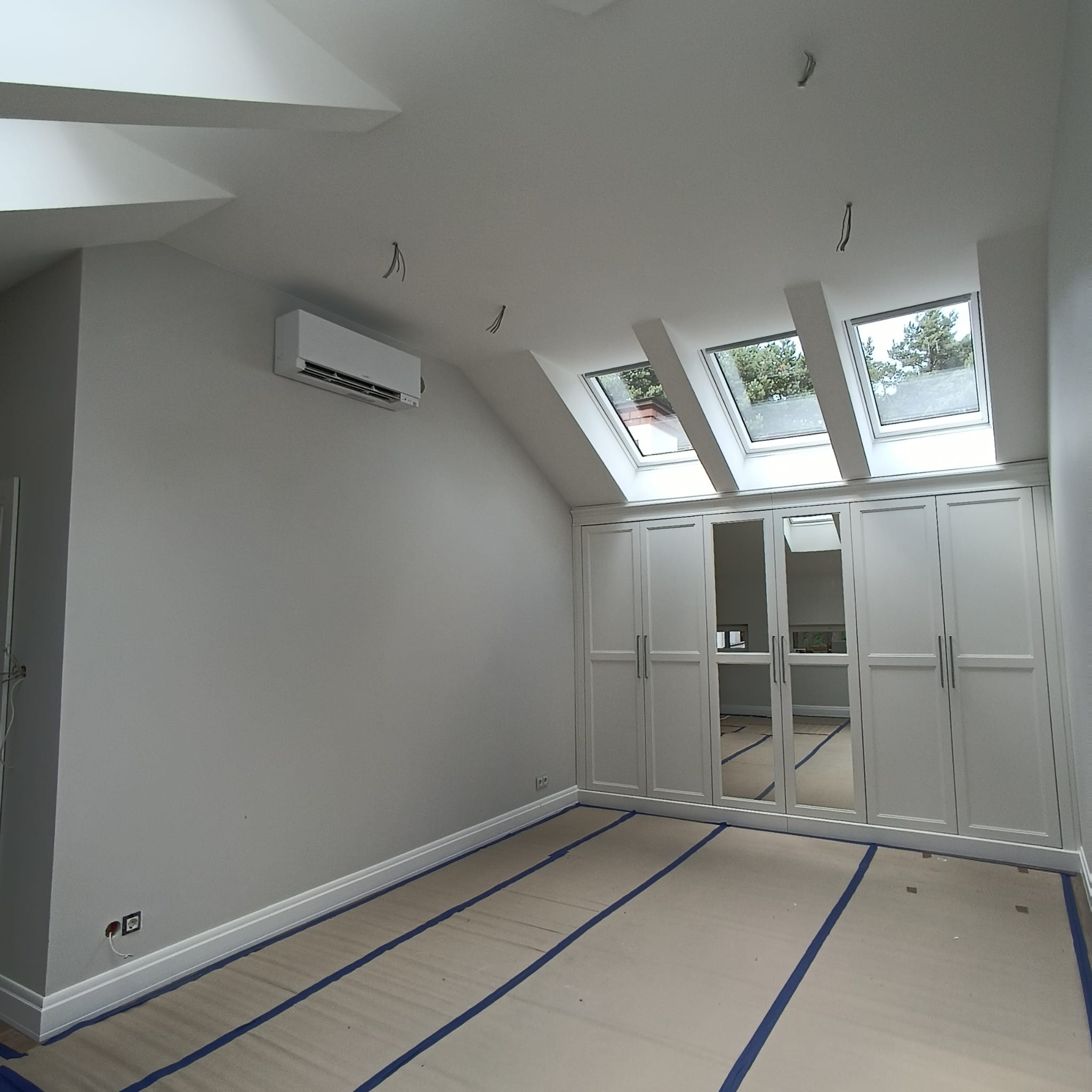 Pokój na poddaszu z trzema oknami dachowymi, zabudowaną szafą z lustrami, klimatyzacją i zabezpieczoną podłogą przed zabrudzeniem podczas prac wykończeniowych.