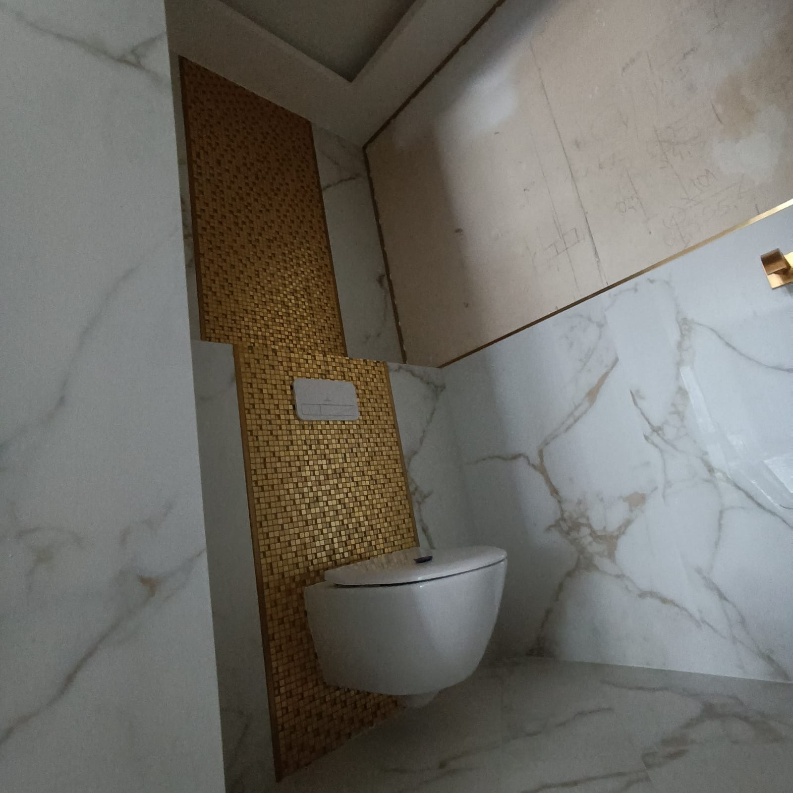 Wisząca toaleta na tle ściany wyłożonej złotą mozaiką w łazience z marmurowymi płytkami, widoczne niedoskonałości na ścianie przed położeniem płytek.