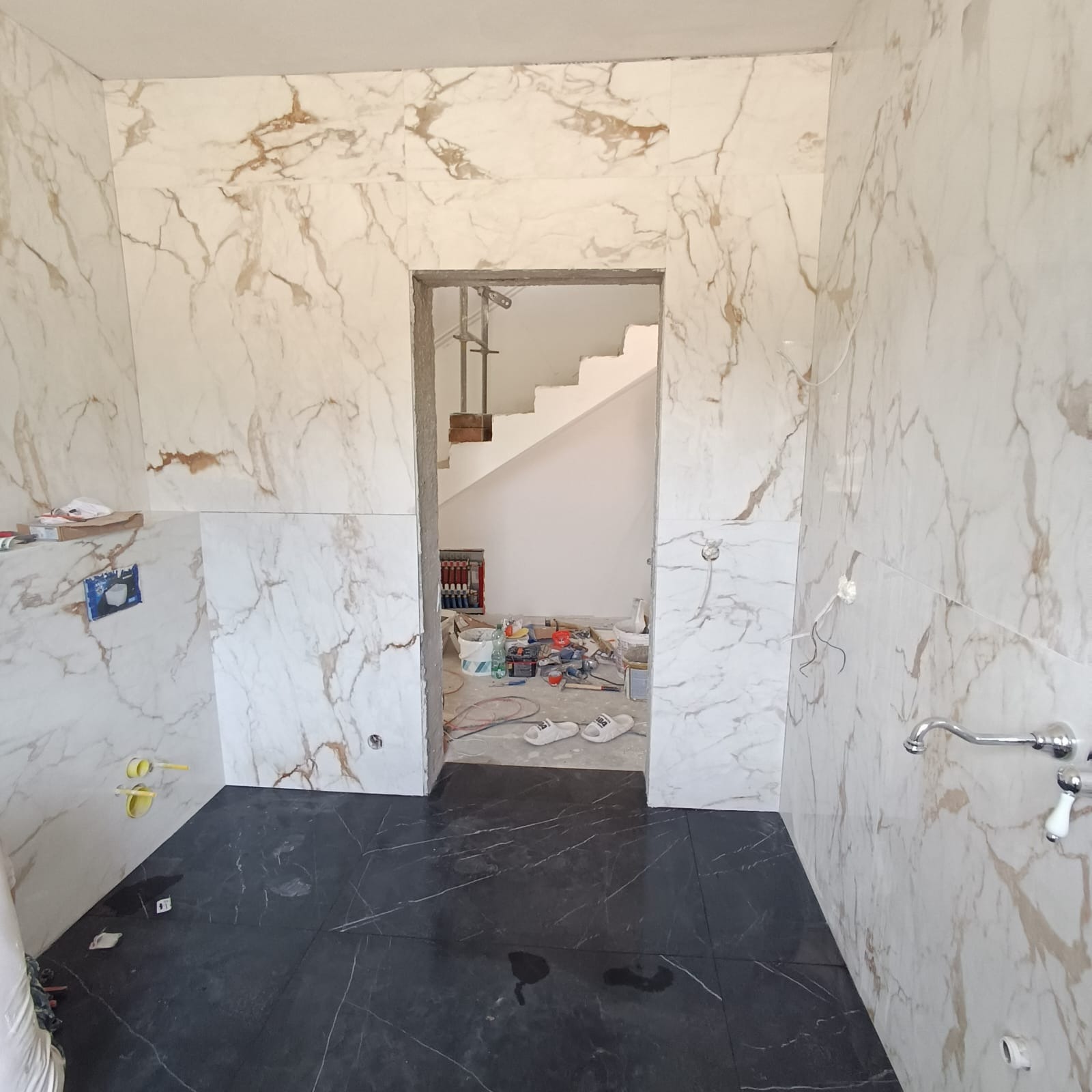 Wnętrze łazienki w trakcie prac wykończeniowych: marmurowe płytki na ścianach, ciemna podłoga, widoczne instalacje, narzędzia i materiały budowlane w przejściu do kolejnego pomieszczenia, gdzie...