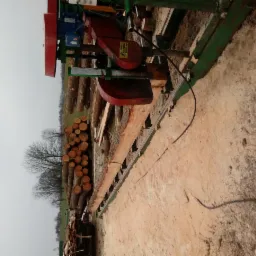 Przenośna traka taśmowa tnie kłodę drewna na deski, widoczne stosy pociętych bali w tle, pochmurny dzień.