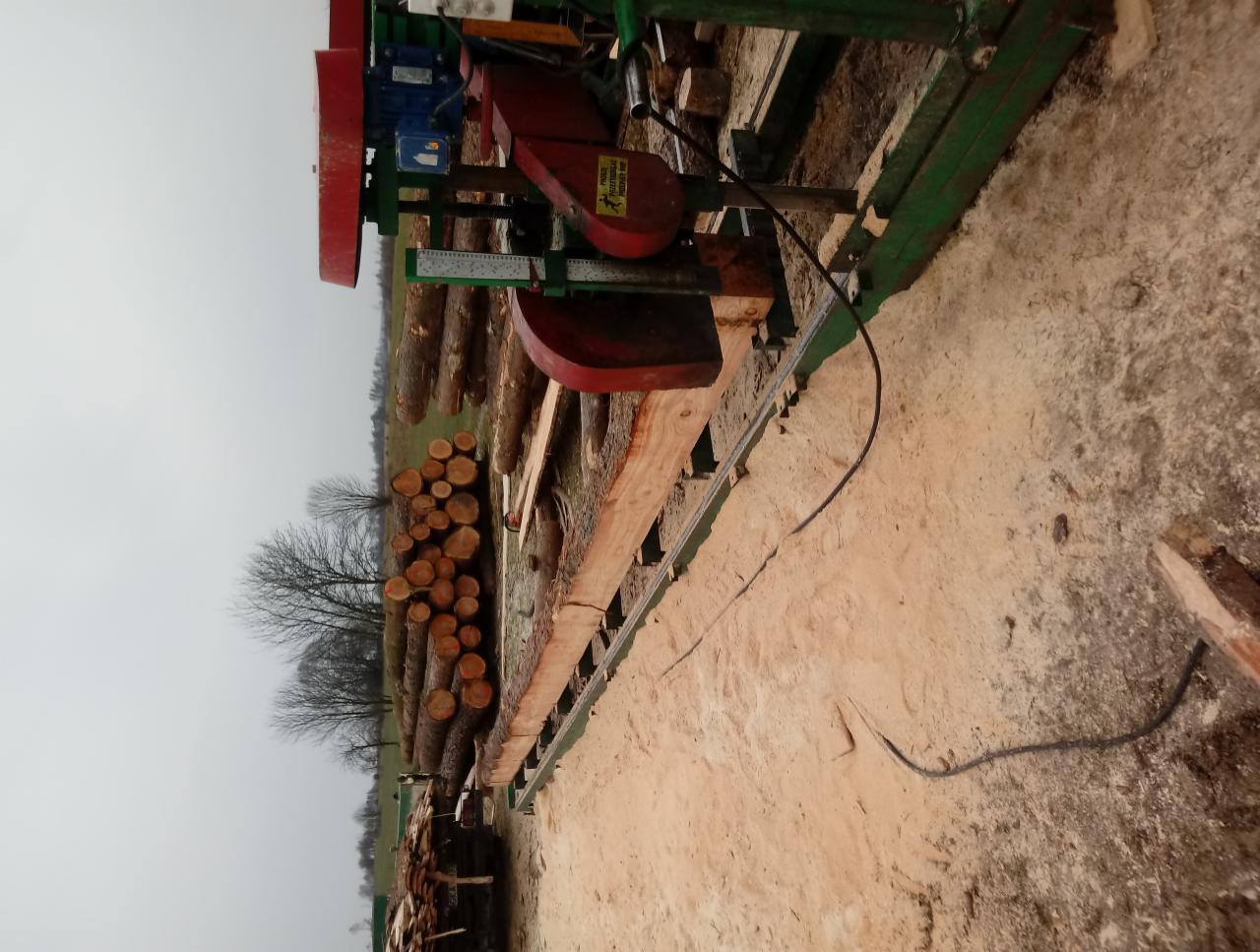 Przenośna traka taśmowa tnie kłodę drewna na deski, widoczne stosy pociętych bali w tle, pochmurny dzień.