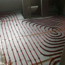 Czerwone rury układane spiralnie na podłodze, mocowane czarnymi uchwytami, przygotowane do zalania wylewką. Widoczne fragmenty folii ochronnej i otwory w ścianie.