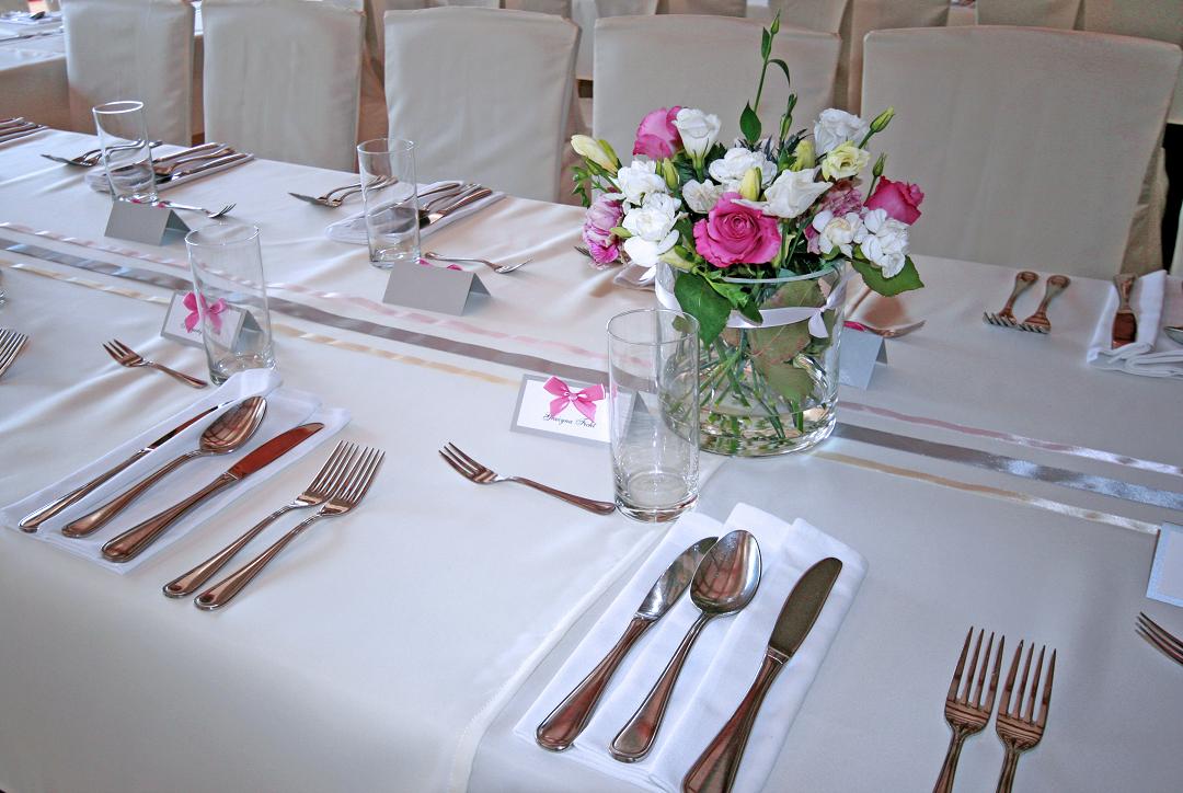 Elegancko nakryty stół z białym obrusem, srebrnymi sztućcami ułożonymi na serwetkach, szklankami, wizytówkami z różowymi kokardkami i bukietem różowych i białych kwiatów w szklanym wazonie.