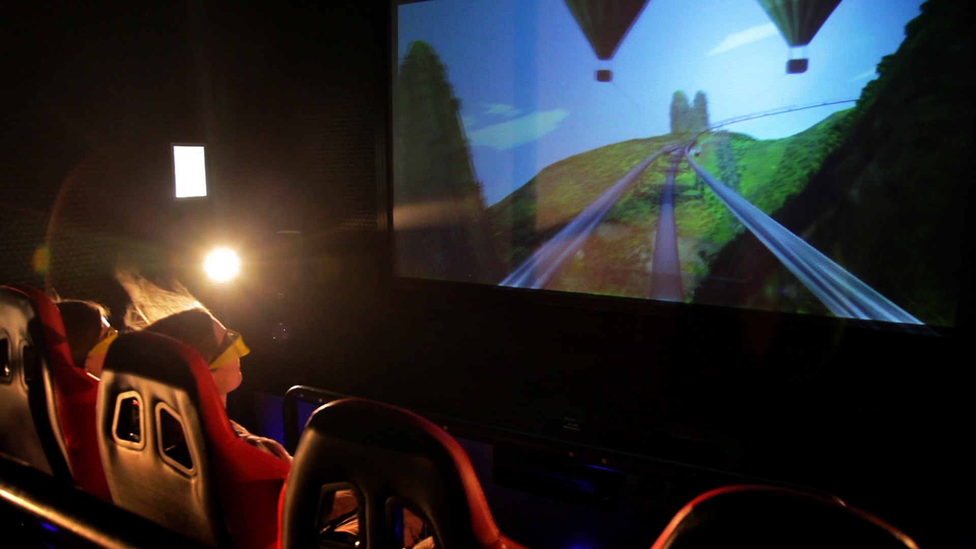 Widok z fotela kinowego na ekran 3D z animacją kolejki górskiej i balonów, widzowie w okularach 3D.