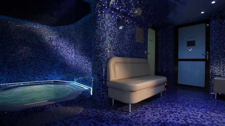 Mozaikowa wanna jacuzzi z podświetleniem LED, obok beżowa sofa w strefie relaksu spa, ściany i podłoga wyłożone niebieską mozaiką.