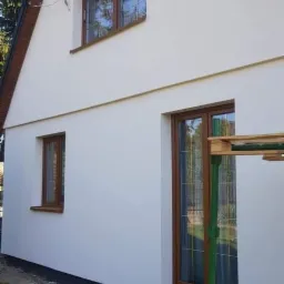 Świeżo otynkowana, jasna elewacja domu jednorodzinnego z brązowymi oknami, widoczne fragmenty rusztowania w kolorze zielonym.