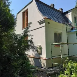 Dwukondygnacyjny dom z nową, jasną elewacją, widoczne rusztowania i fragment ogrodu, w tle błękitne niebo.