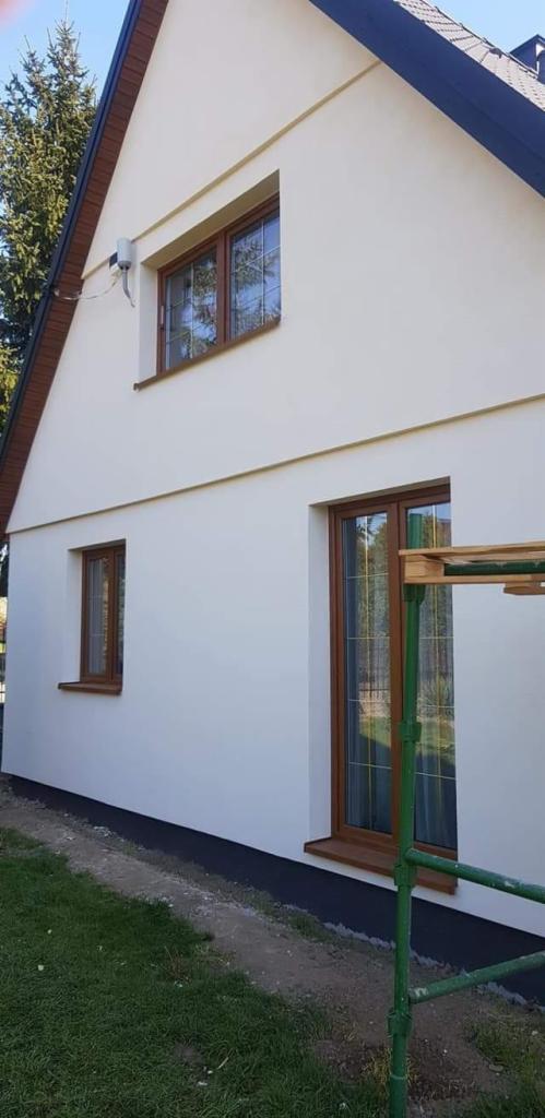 Świeżo otynkowana, jasna elewacja domu jednorodzinnego z brązowymi oknami, widoczne fragmenty rusztowania w kolorze zielonym.