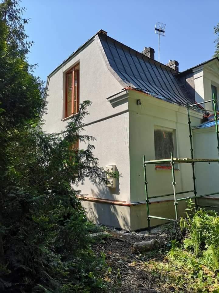 Dwukondygnacyjny dom z nową, jasną elewacją, widoczne rusztowania i fragment ogrodu, w tle błękitne niebo.