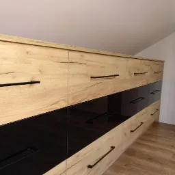 Długa komoda z szufladami, fronty z drewna w kolorze jasnego dębu i czarnego szkła, podłużne czarne uchwyty, zamontowana na poddaszu ze skosem.