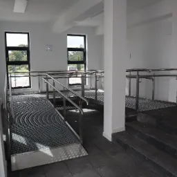 Wewnętrzny podjazd dla wózków inwalidzkich z metalową kratką i poręczami ze stali nierdzewnej, widok z góry na schody i okna z widokiem na zewnątrz, w tle zaparkowany samochód.