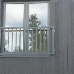 Szara balustrada okienna z metalowymi pionowymi prętami, zamontowana na elewacji budynku z szarego drewna, okno z odbijającym się drzewem.