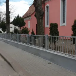 Szara metalowa barierka z motywem ryb, zamontowana na niskim betonowym murku wzdłuż chodnika przed różowym budynkiem. Na pierwszym planie zaparkowany szary samochód osobowy.