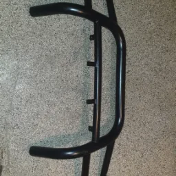 Czarna, metalowa konstrukcja ochronna z rur, prawdopodobnie orurowanie lub element ramy, leżąca na szarej, teksturowanej powierzchni.