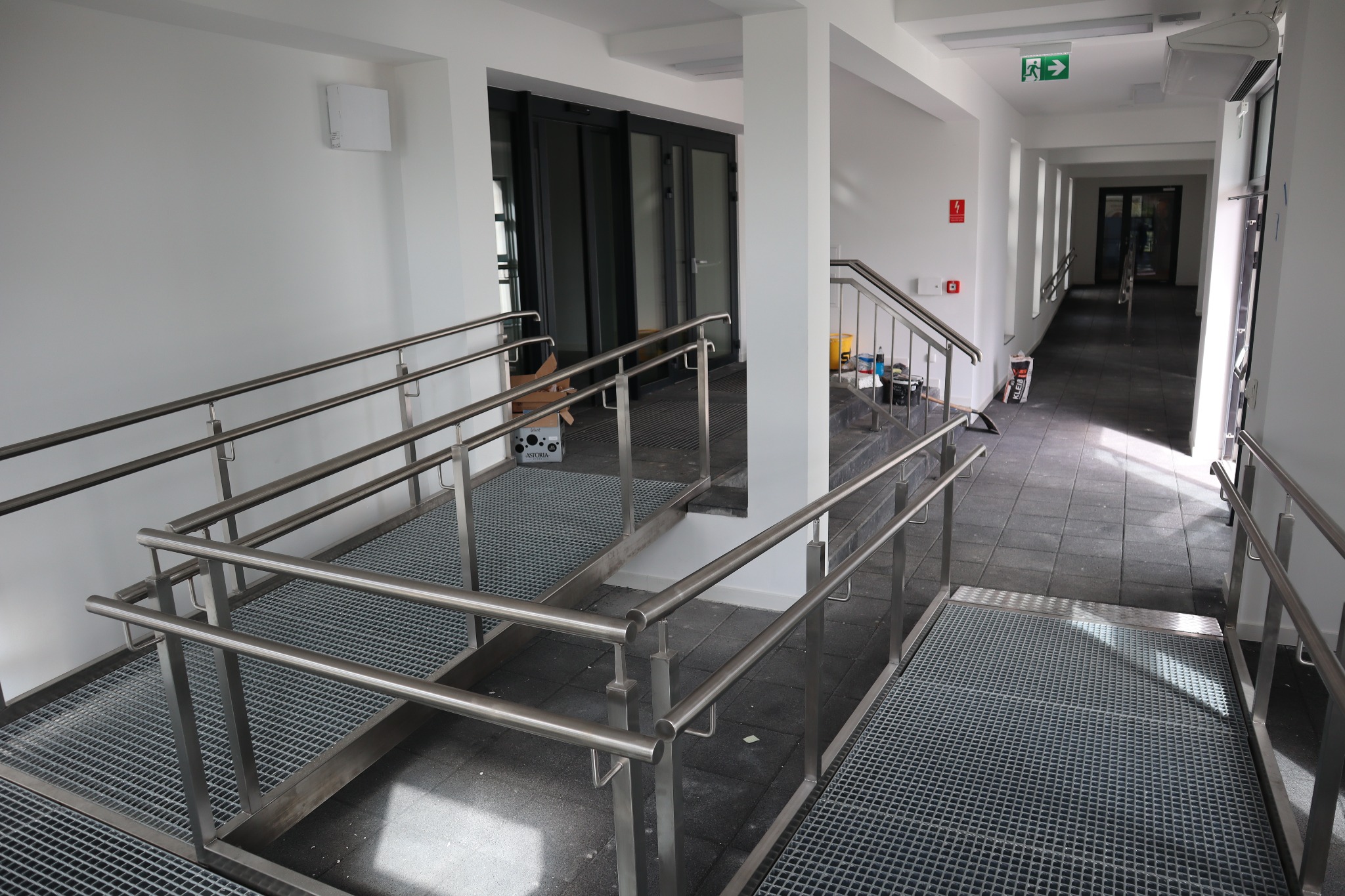 Wewnętrzny korytarz z podjazdem dla wózków inwalidzkich wykonanym z metalowych krat i balustrad ze stali nierdzewnej, widoczne drzwi wejściowe i schody.