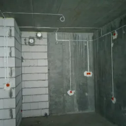 Surowy betonowy pokój z widocznymi przewodami elektrycznymi w białych rurkach, wystającymi ze ścian i sufitu, z pomarańczowymi puszkami instalacyjnymi, widoczne ślady po wierceniu w ścianach.