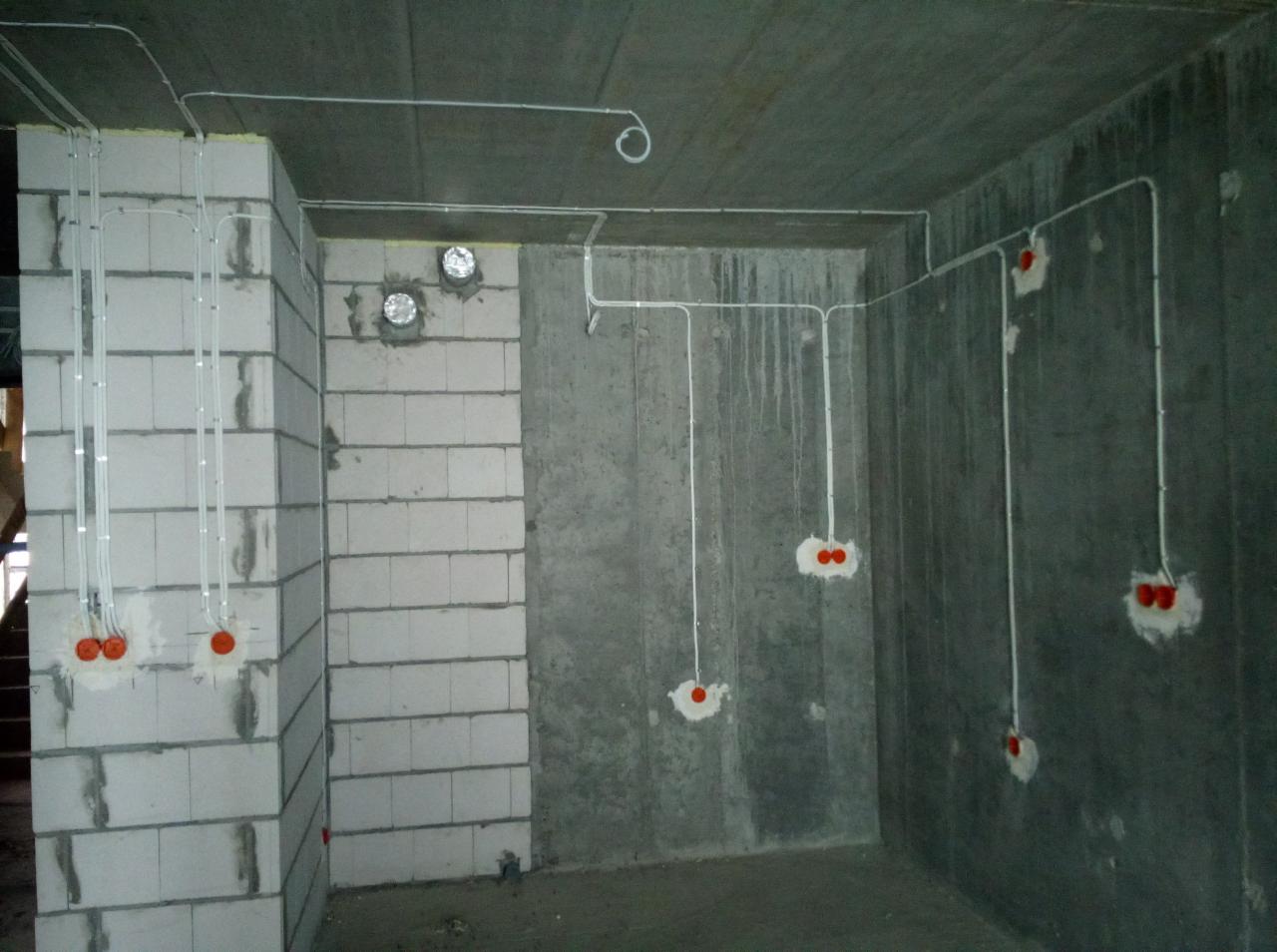 Surowy betonowy pokój z widocznymi przewodami elektrycznymi w białych rurkach, wystającymi ze ścian i sufitu, z pomarańczowymi puszkami instalacyjnymi, widoczne ślady po wierceniu w ścianach.