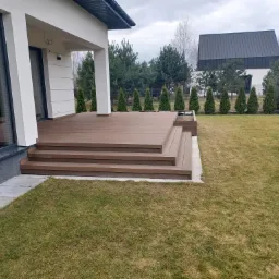 Nowoczesny taras z deski kompozytowej w odcieniach brązu, z trzema stopniami, przylegający do białego domu z dużym oknem, otoczony trawnikiem i tujami.