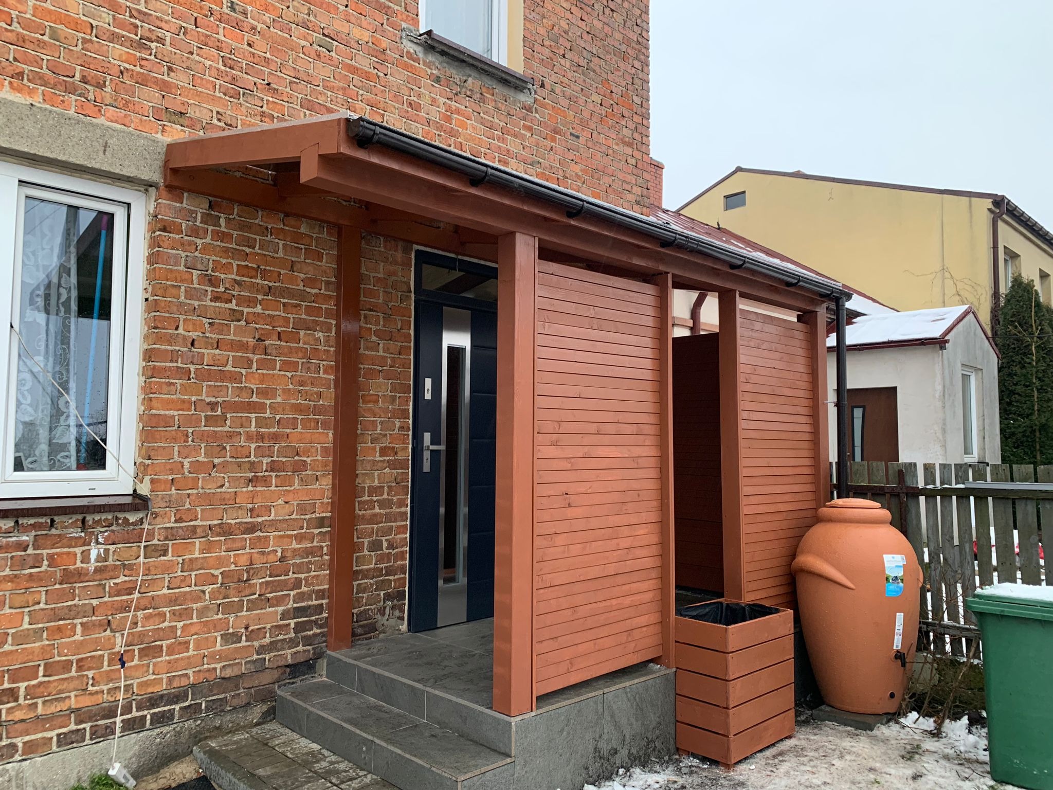 Brązowa drewniana konstrukcja z zadaszeniem przy wejściu do domu z cegły, widoczny zbiornik na deszczówkę i donica.