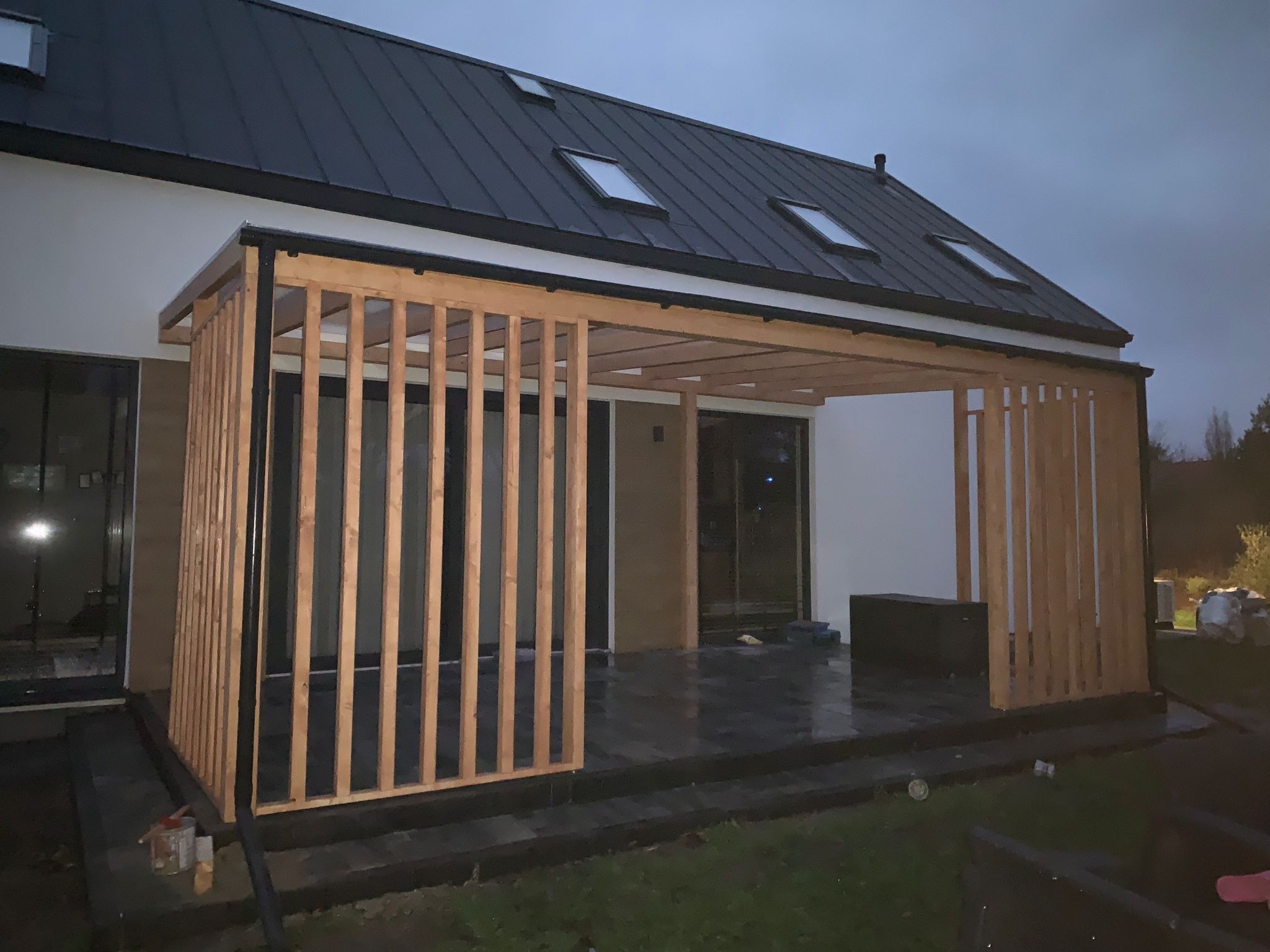 Nowoczesna pergola drewniana z pionowymi listwami, przylegająca do białego budynku z czarnym dachem i oknami dachowymi, widoczna z zewnątrz na tle pochmurnego nieba.