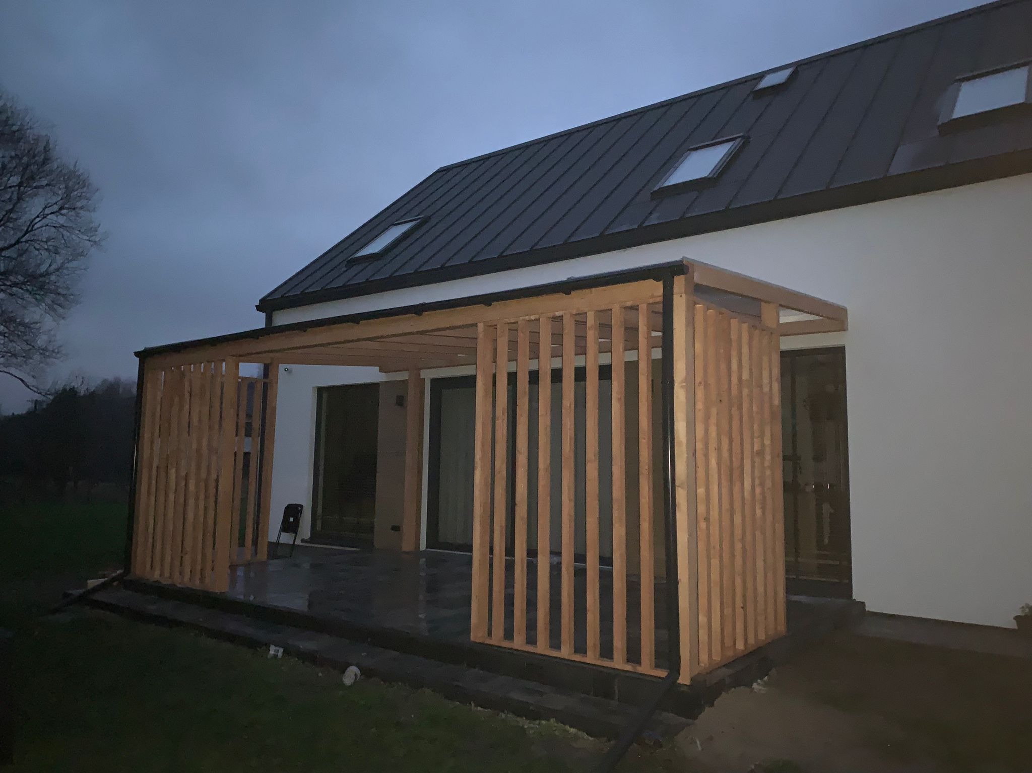 Drewniana konstrukcja pergoli z pionowymi listwami, przylegająca do nowoczesnego domu z ciemnym dachem i oknami dachowymi, widok z zewnątrz w pochmurny dzień.