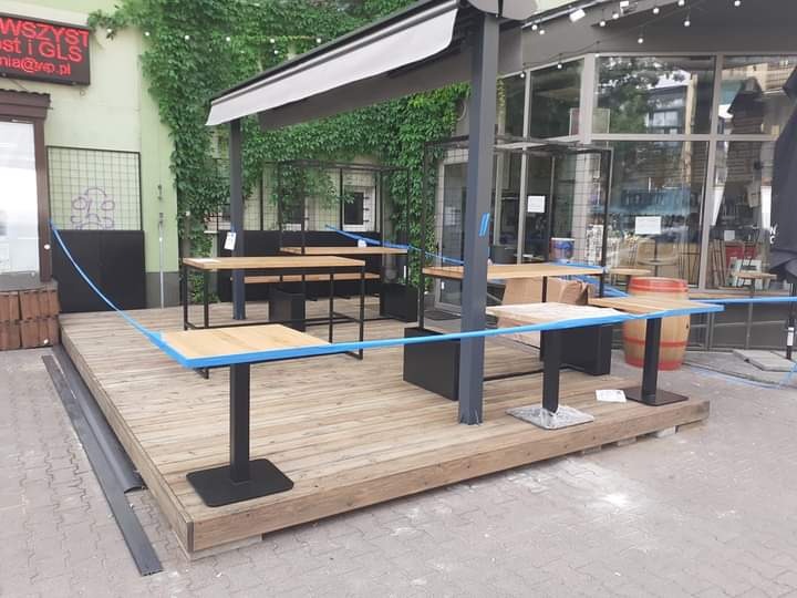 Drewniany taras komercyjny z czarną metalową pergolą i stolikami z jasnego drewna z niebieskimi krawędziami, otoczony niebieską taśmą ostrzegawczą.