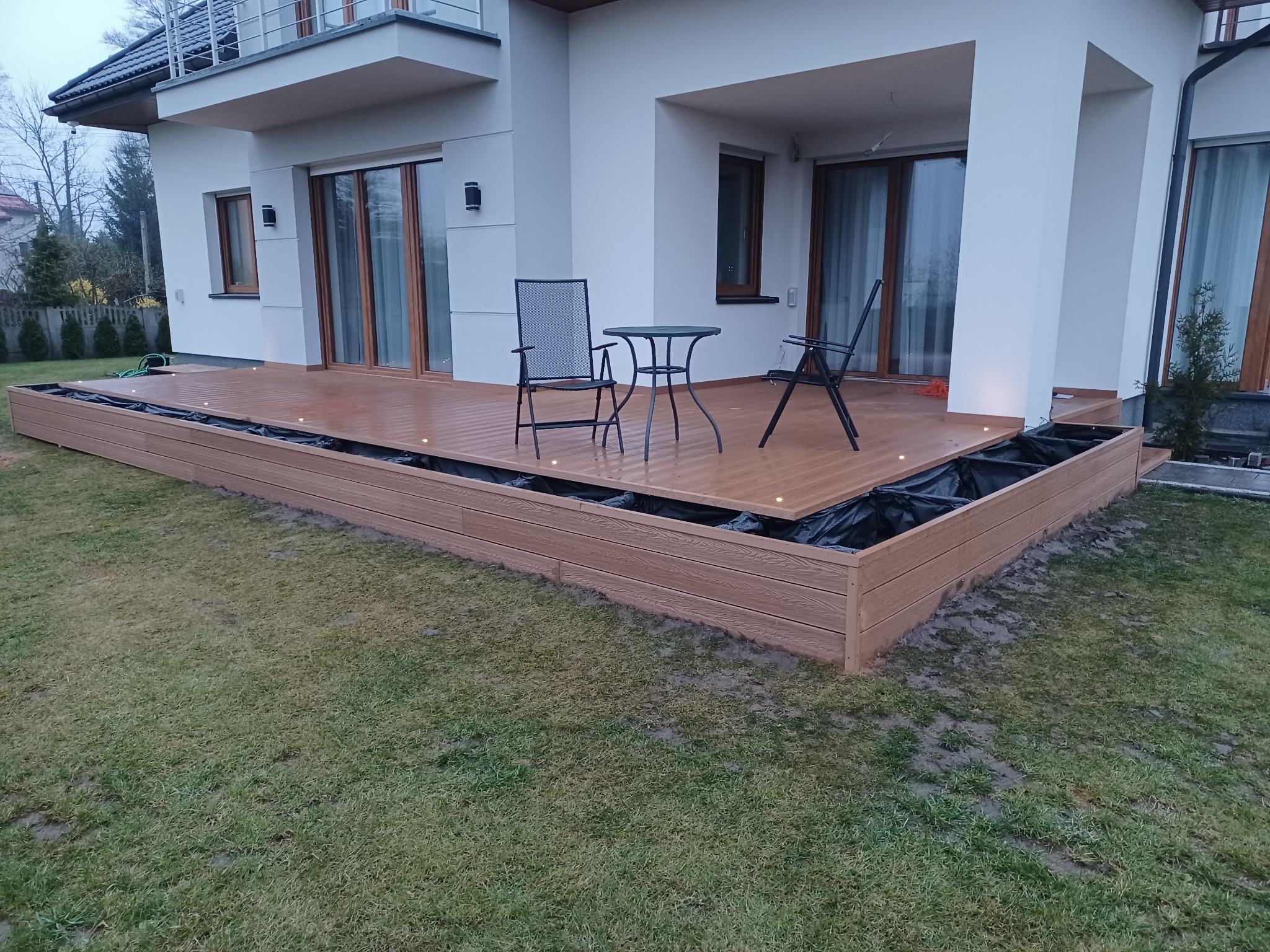 Nowoczesny taras z desek kompozytowych w trakcie montażu, widoczne podświetlenie LED, czarna folia izolacyjna, metalowe krzesła i stolik na tle białego domu z balkonem.